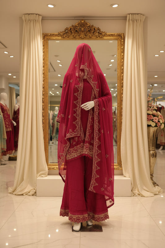 A traditional pink shalwar kameez suit with a matching sheer dupatta and white embroidered borders, standing in front of a patterned curtain.